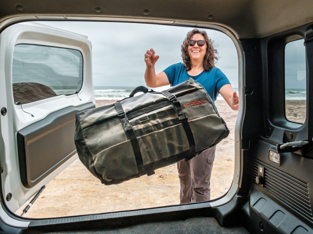 Woman loading a Turkana TurtleTrunk Double-Roll Stuff Bag into a 4x4 vehicle on a beach, showing durable overland and outdoor lifestyle gear.