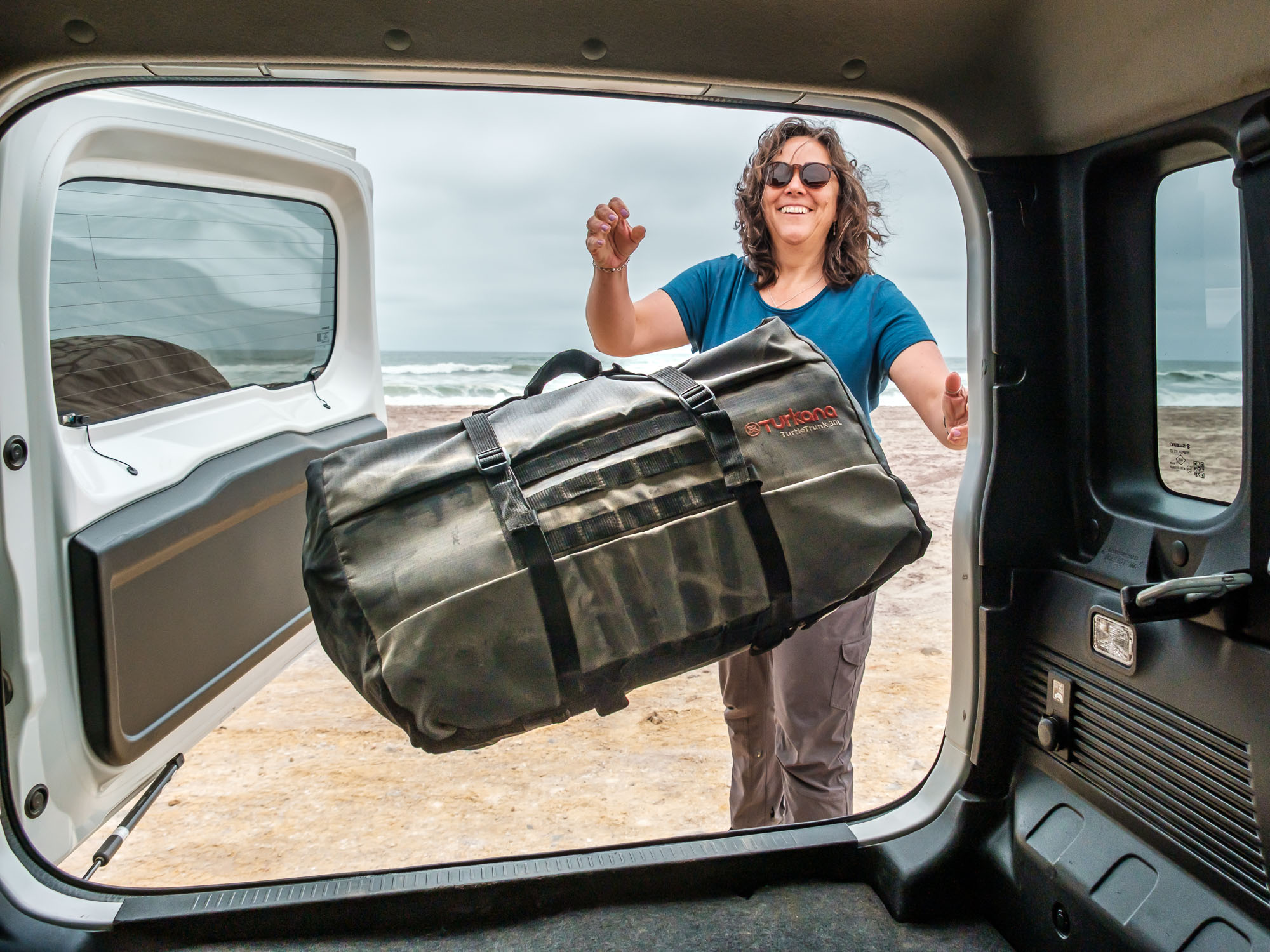 Woman loading a Turkana TurtleTrunk Double-Roll Stuff Bag into a 4x4 vehicle on a beach, showing durable overland and outdoor lifestyle gear.