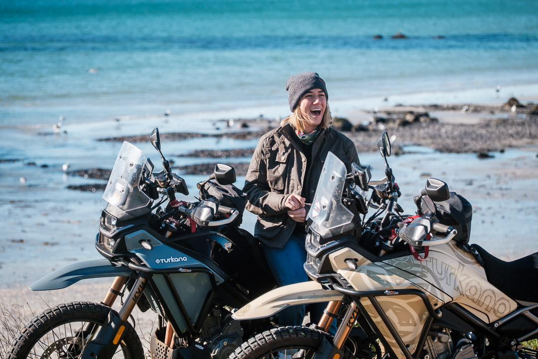 Female adventure motorcycle rider wearing a grey Turkana Nutshell beanie while sitting between two adventure bikes beside the ocean.