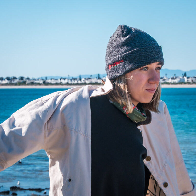 Close-up of female motorcycle traveller wearing a grey Turkana beanie beside adventure motorcycles near the shoreline.