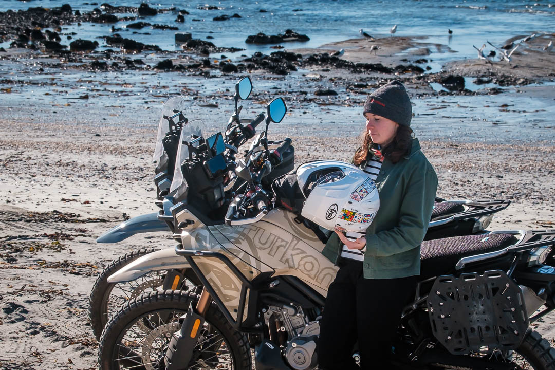 Close-up of female motorcycle traveller wearing a grey Turkana beanie beside adventure motorcycles near the shoreline.
