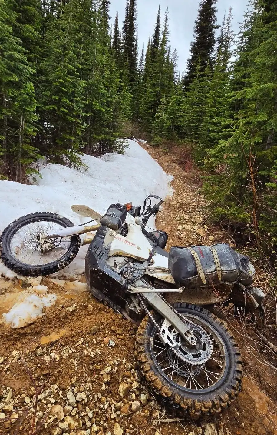 Mud-covered adventure motorcycle with Turkana soft luggage lying on a rocky forest trail beside melting snow.
